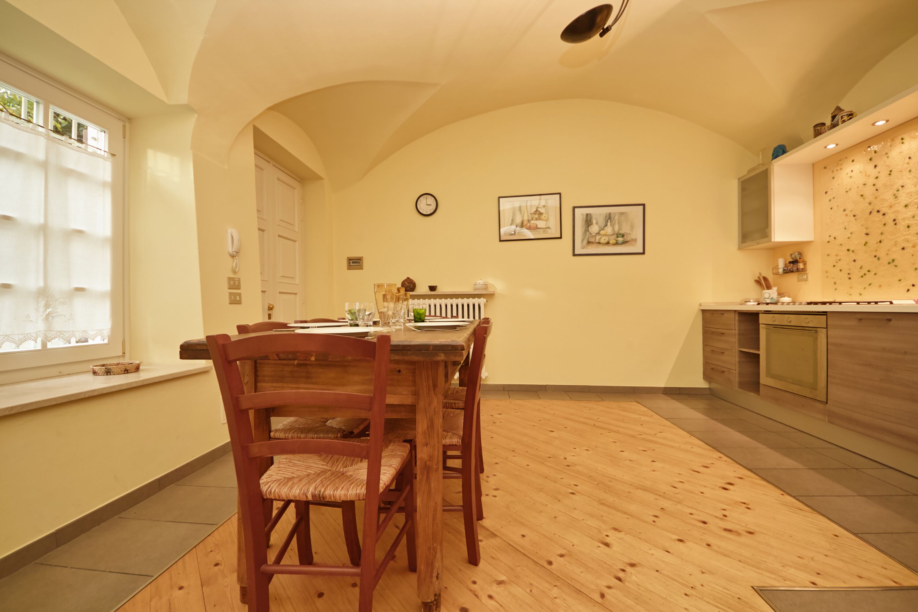 Un comedor con una mesa y sillas de madera, un reloj en la pared y una cocina al fondo.