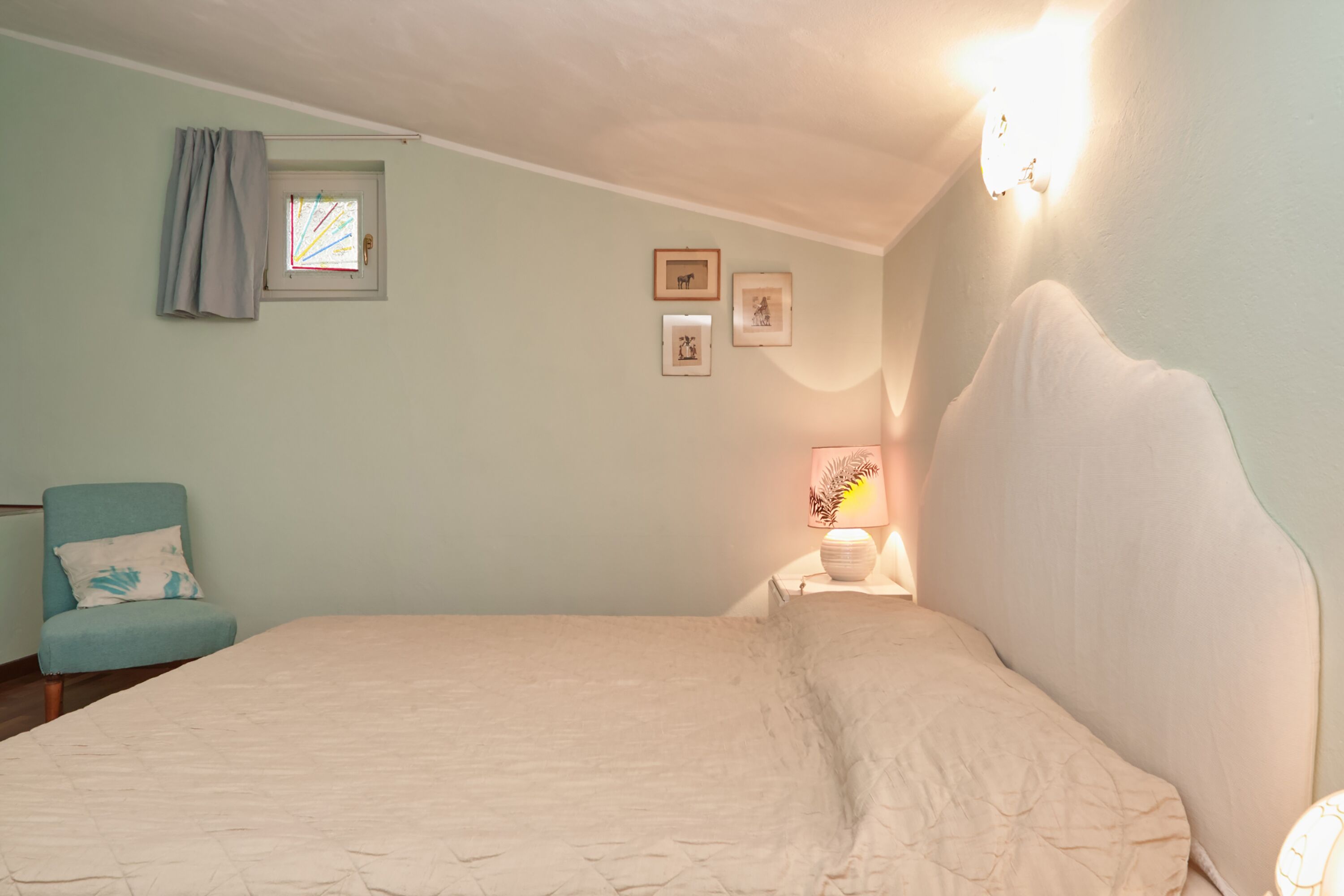 A bedroom with a bed, a chair, a nightstand with a lamp, and framed pictures on the wall.