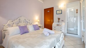 A bedroom with a large bed, a wooden door, a white dresser, and a bathroom visible through a glass door. - Le Notti Mediterranee (Cervo)
