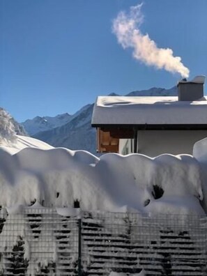 A building with a snow-covered roof and a chimney emitting smoke, surrounded by a mountainous landscape. - Haus Hubertus (Jerzens)
