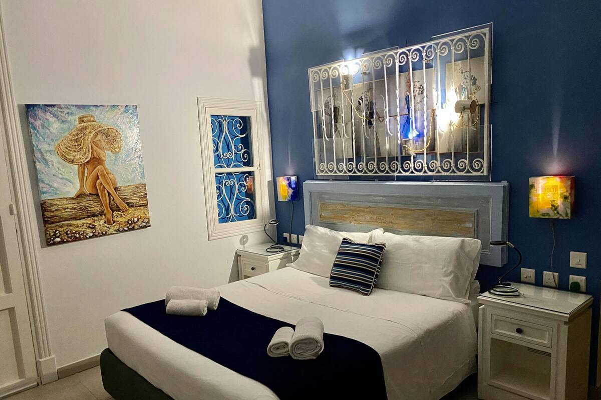 A neatly made bed with white and navy bedding, two bedside tables with lamps, a wall-mounted TV, and artwork on the walls.
