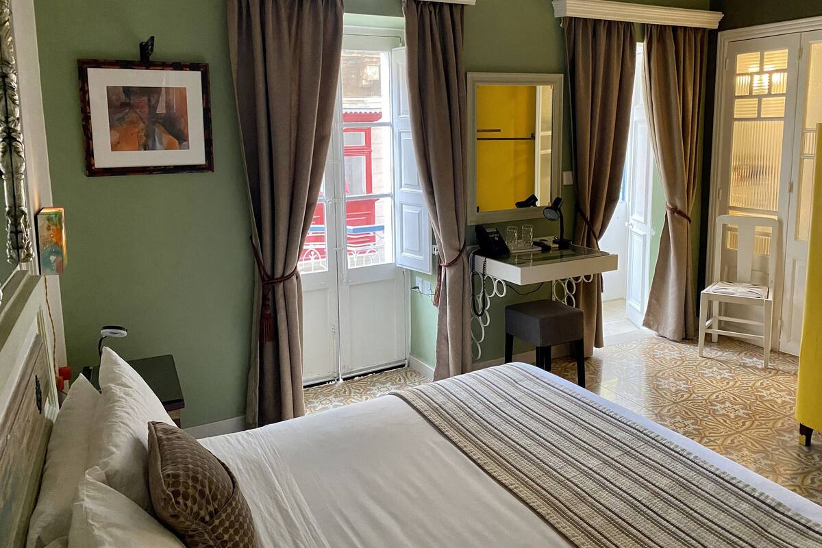 A hotel room with a bed, a desk with a telephone and glasses, a mirror, and a door leading to a balcony.