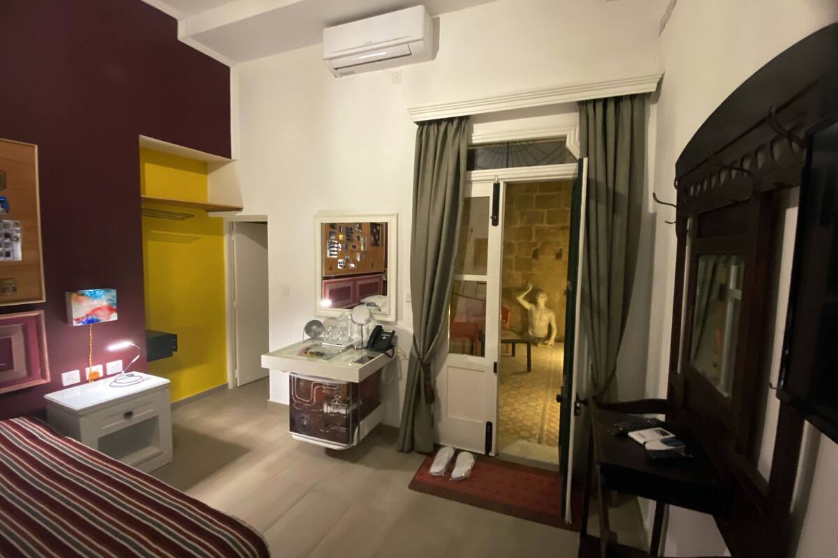 A hotel room with a bed, bedside table, lamp, and a small desk with a chair. There is a door leading to a bathroom and a window with curtains.