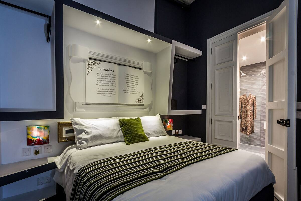 A modern bedroom with a large bed, a nightstand, a wall-mounted reading light, and a door leading to another room.