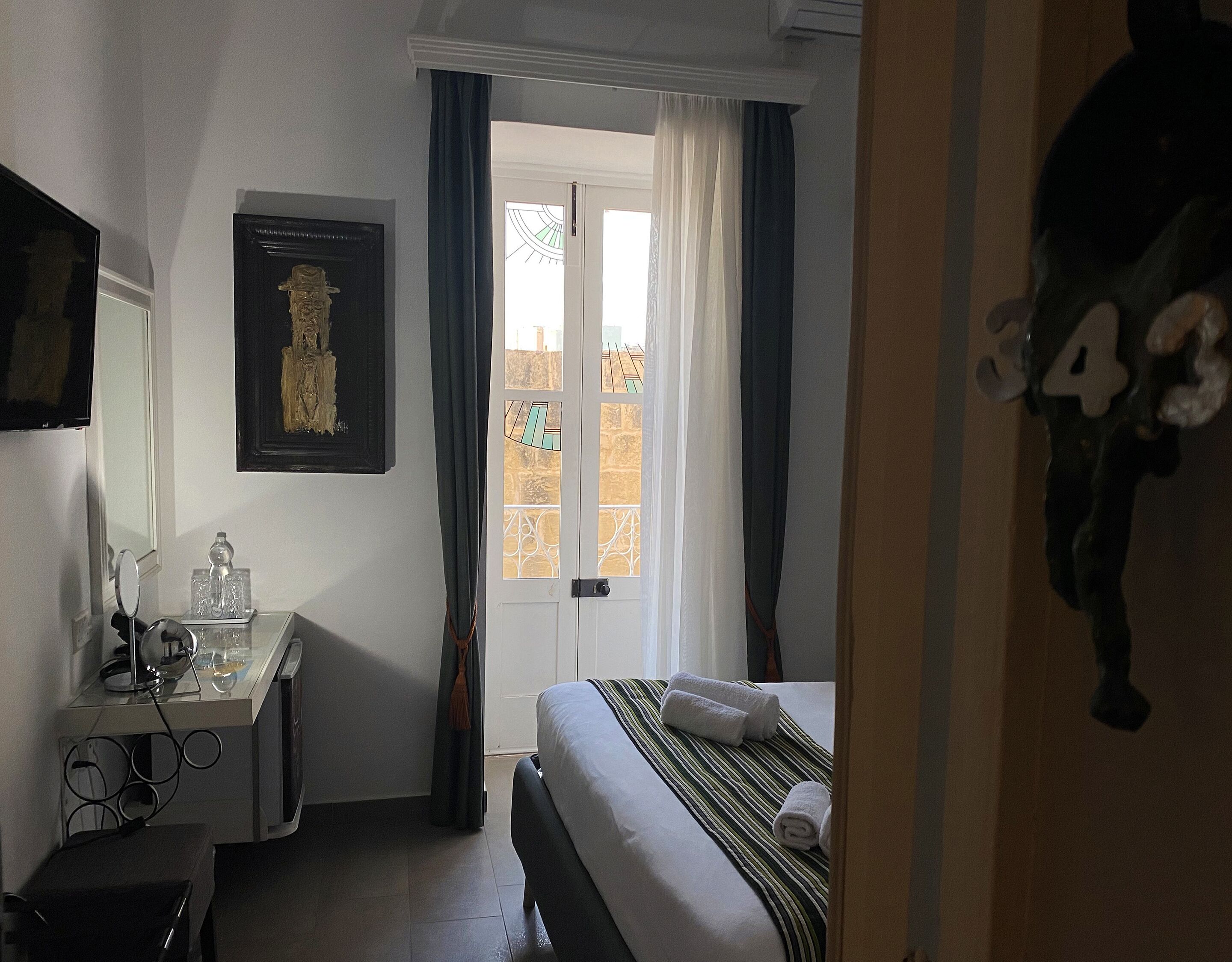 A hotel room with a bed, a desk with a mirror, a framed artwork, and a window with curtains.