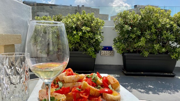 A table with a plate of bruschetta and a glass of white wine, surrounded by potted plants. - BOCO Boutique (Cospicua)