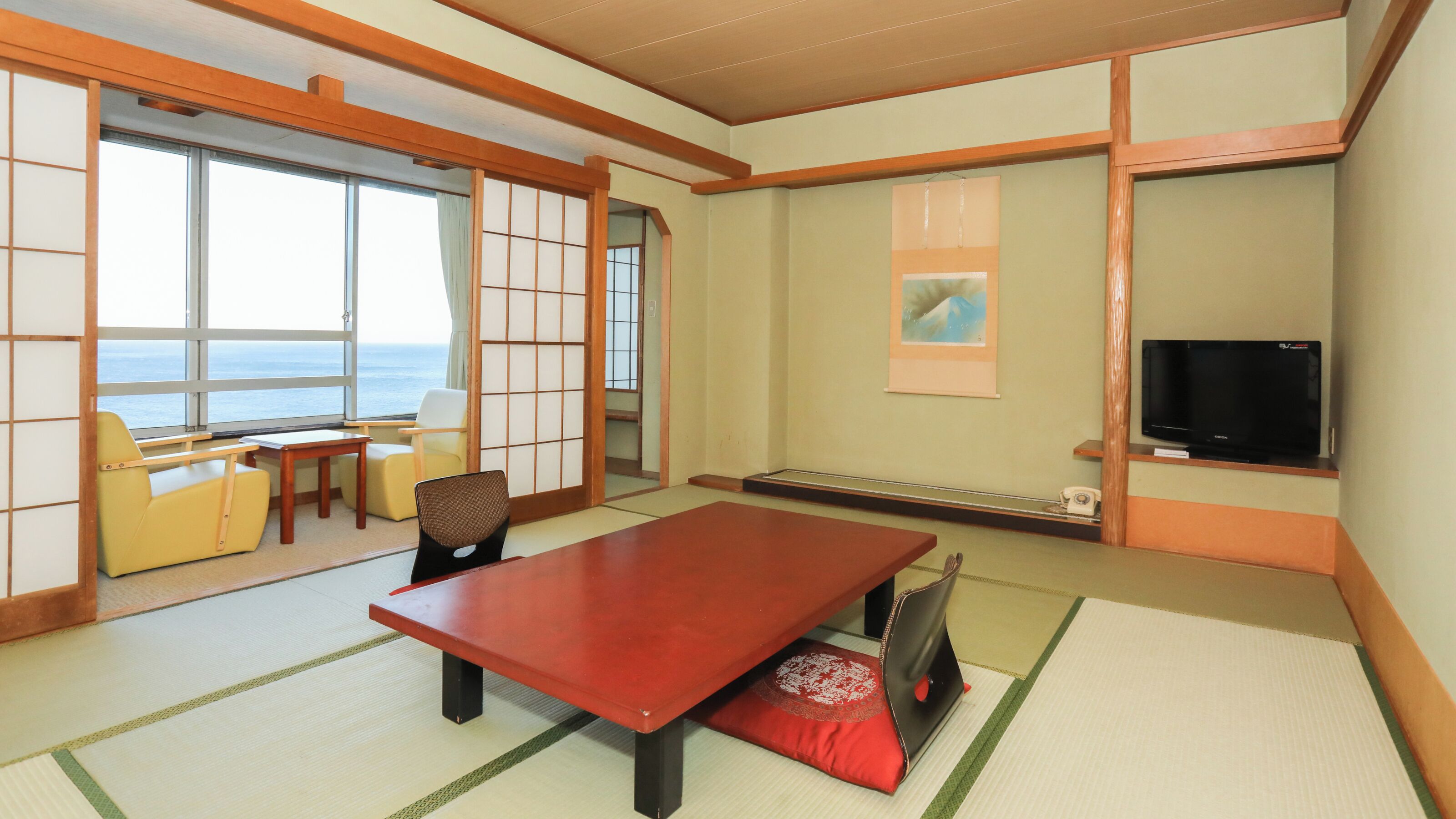 A traditional Japanese room with tatami flooring, a low table, chairs, a television, and a view of the sea.