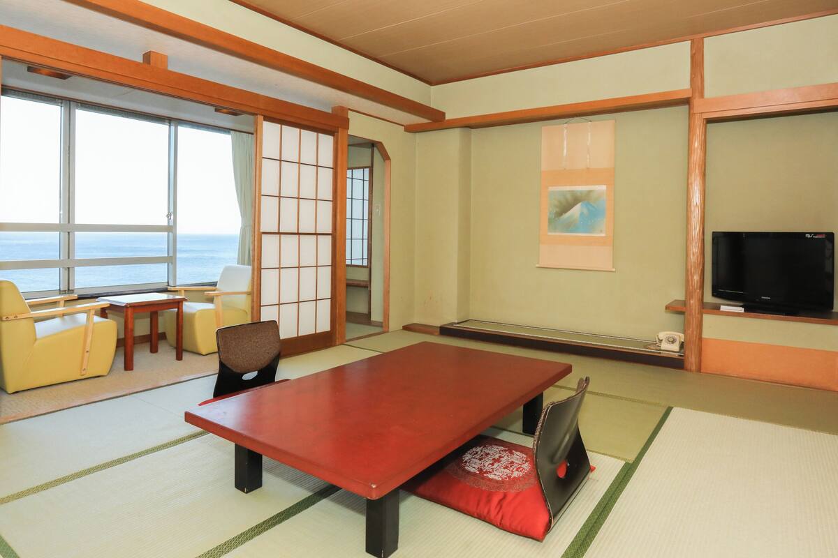 Ein traditioneller japanischer Raum mit Tatami-Böden, einem niedrigen Tisch, Stühlen, einem Fernseher und Meerblick.