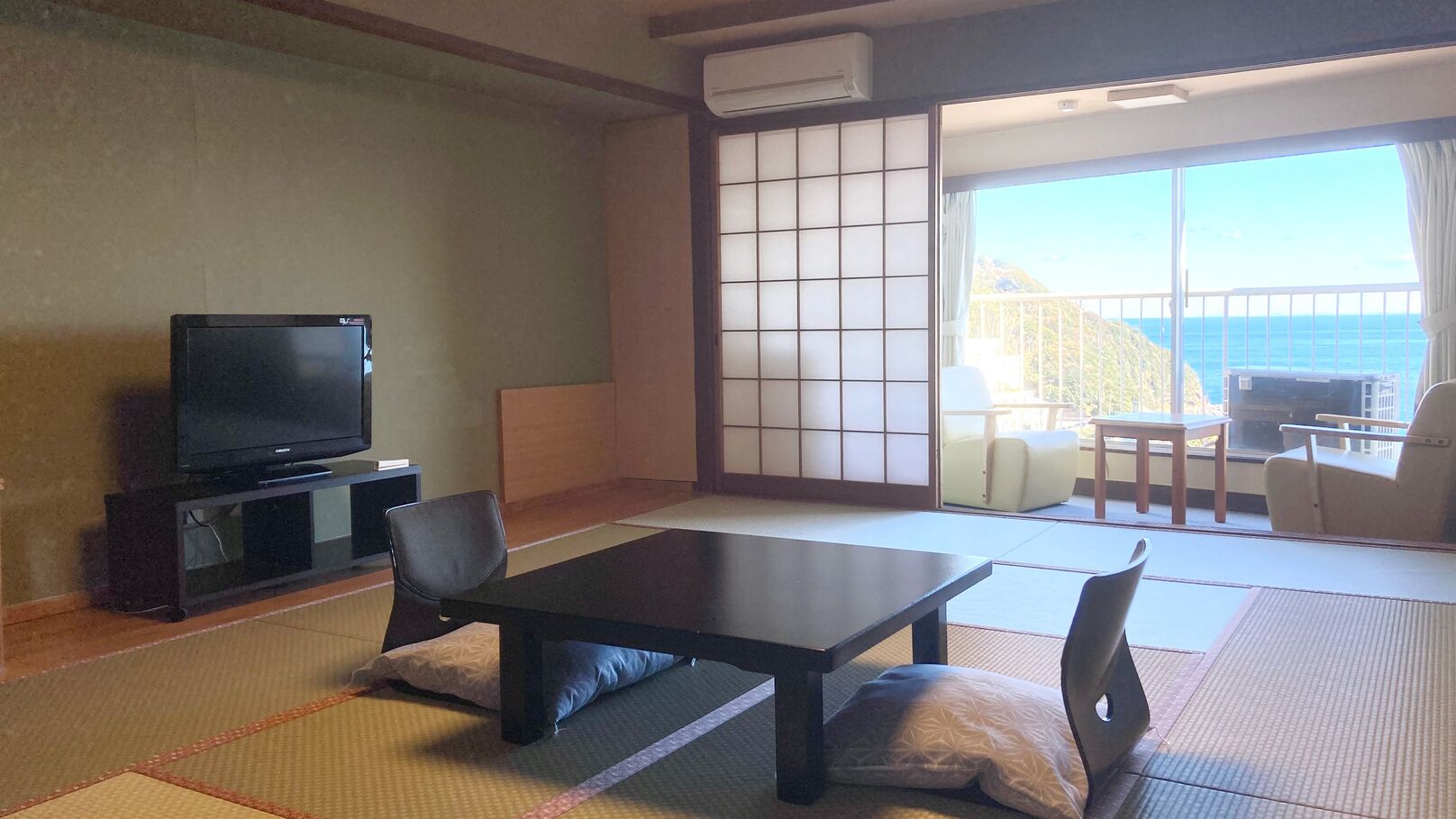 A room with tatami flooring, a television, a sliding door to a balcony, and a view of the ocean.