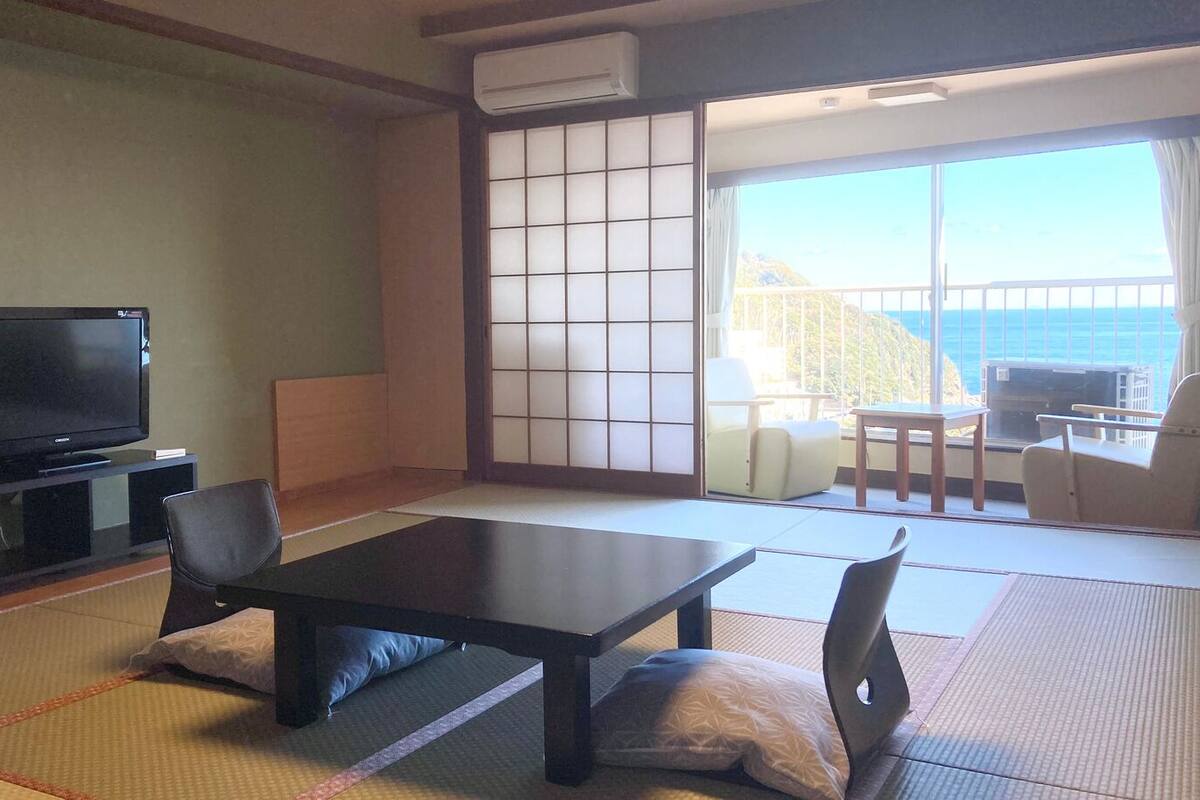Ein Zimmer mit Tatami-Böden, einem Fernseher, einer Schiebetür zum Balkon und Blick auf das Meer.