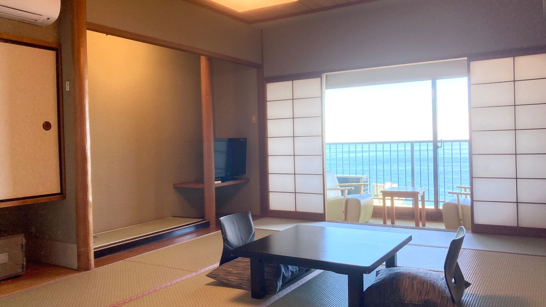 A room with tatami flooring, a low table, chairs, a television on a shelf, and a balcony with a view.