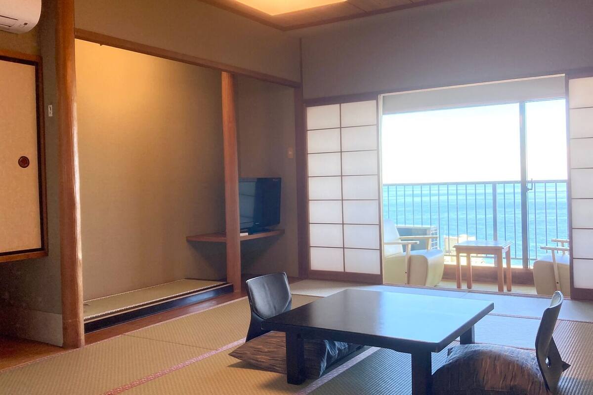 Ein Zimmer mit Tatami-Boden, einem niedrigen Tisch, Stühlen, einem Fernseher auf einem Regal und einem Balkon mit Blick.