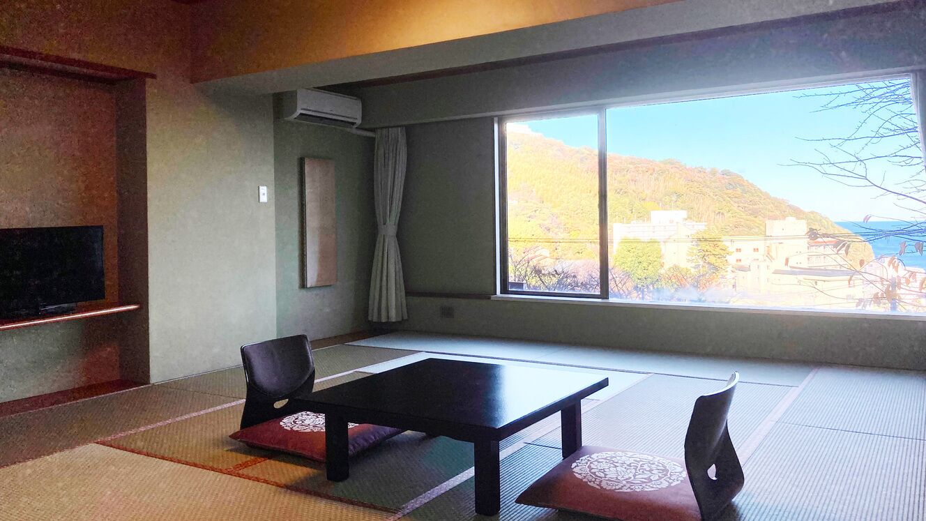 A room with tatami mats, a black table, two chairs, a TV, and a large window with a view of a hillside.