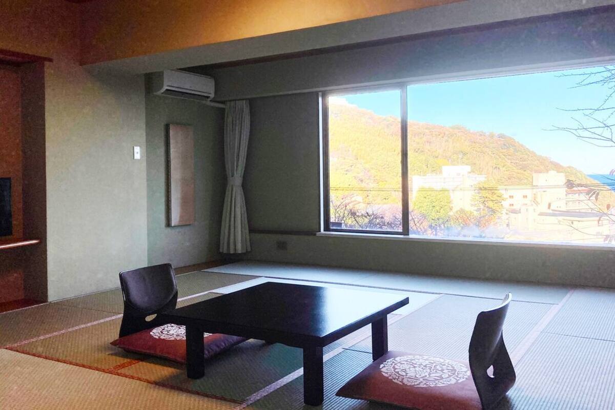 Ein Zimmer mit Tatami-Matten, einem schwarzen Tisch, zwei Stühlen, einem Fernseher und einem großen Fenster mit Blick auf einen Hügel.