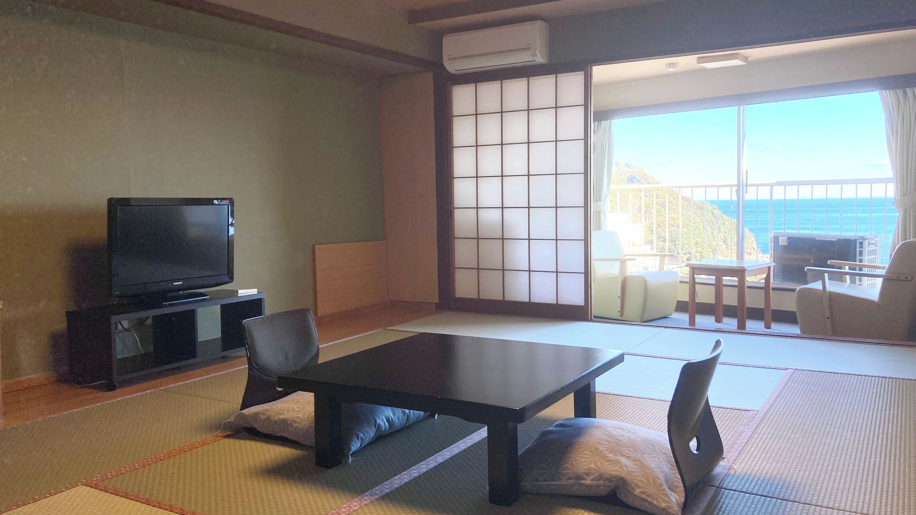 A traditional Japanese-style room with tatami flooring, a low table, chairs, a television, and a sliding glass door leading to a balcony with a view of the sea.