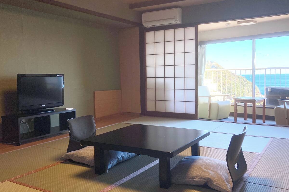 Ein traditioneller japanischer Raum mit Tatami-Boden, einem niedrigen Tisch, Stühlen, einem Fernseher und einer Glastür, die auf einen Balkon mit Meerblick führt.