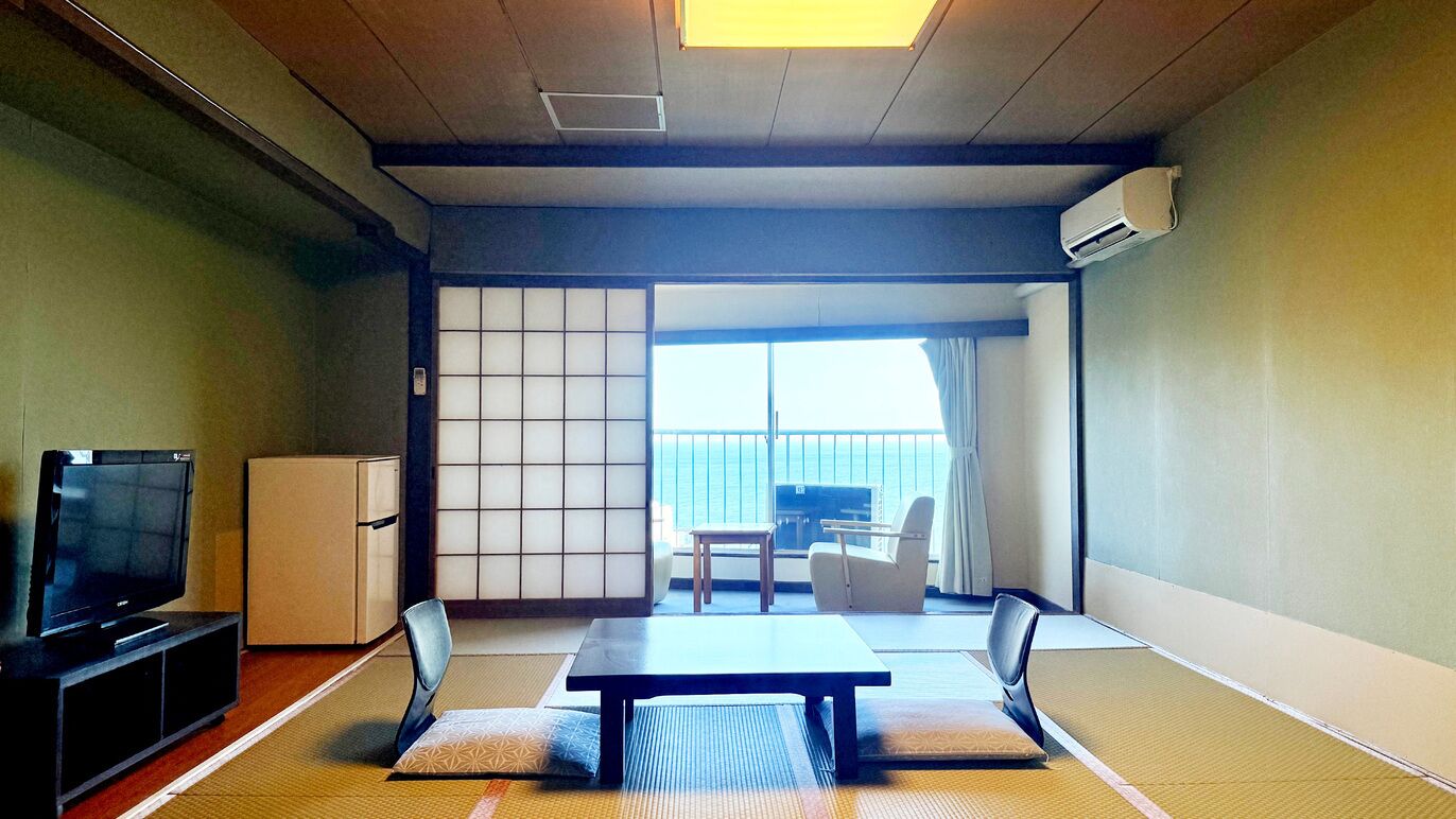 A traditional Japanese room with tatami flooring, a television, a small refrigerator, a sliding door leading to a balcony with a view of the sea.