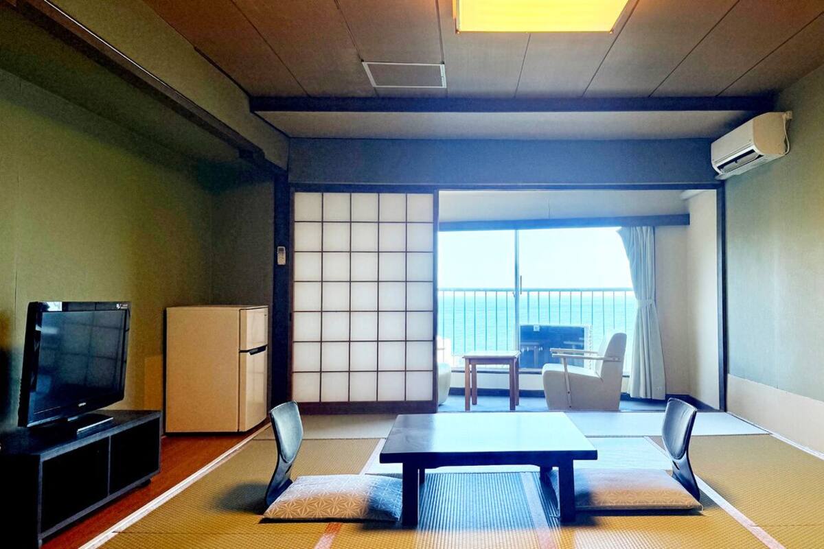 Ein traditioneller japanischer Raum mit Tatami-Böden, einem Fernseher, einem kleinen Kühlschrank und einer Schiebetür zu einem Balkon mit Meerblick.