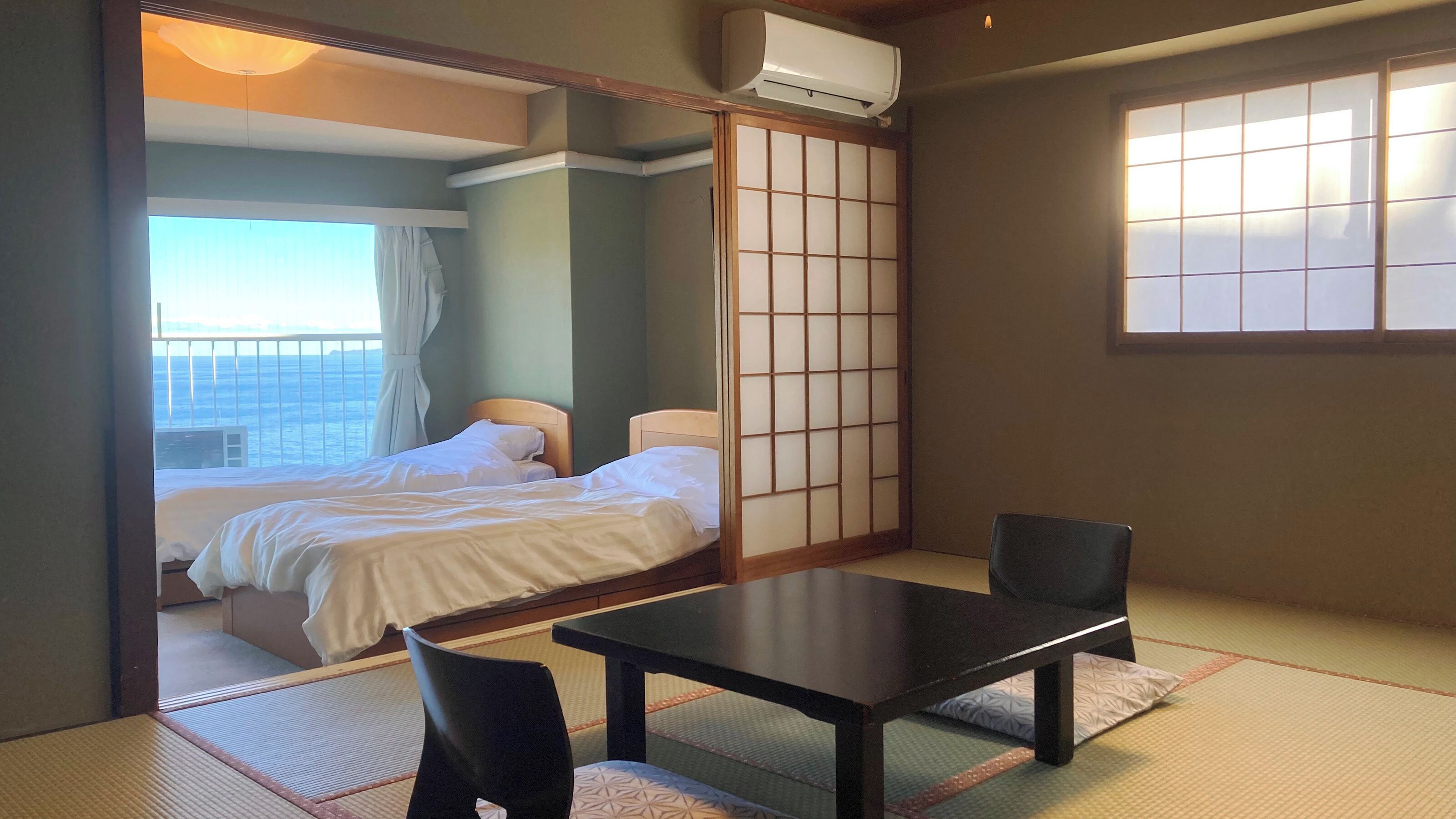A hotel room with a bed, a table, a chair, a sliding shoji screen, and a view of the sea.