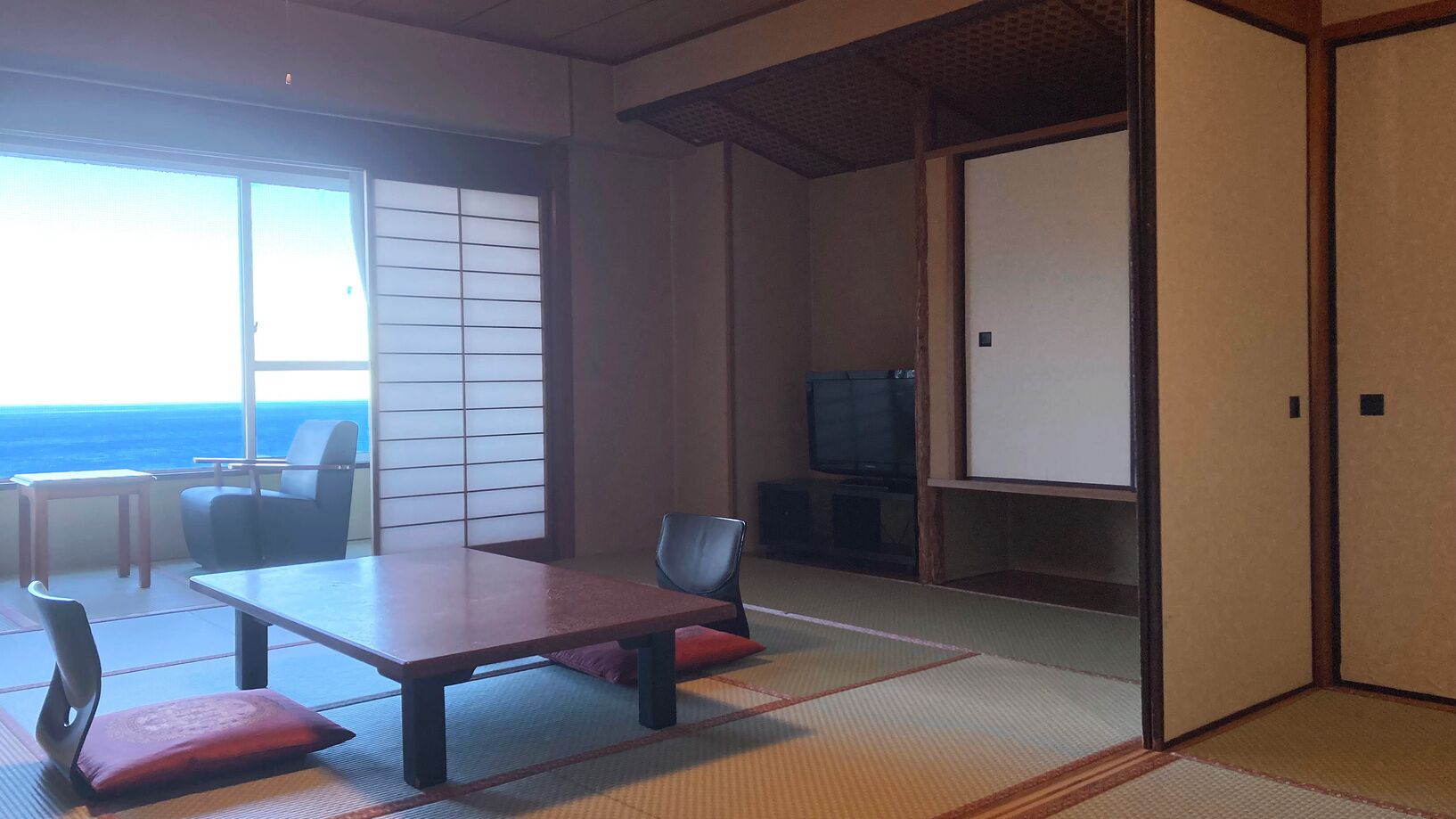 A traditional Japanese room with tatami flooring, a low table with cushions, a television, and a sliding door leading to a balcony with a view of the sea.