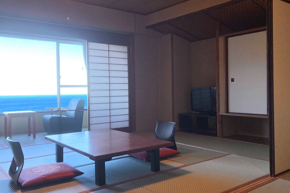 Ein traditioneller japanischer Raum mit Tatami-Böden, einem niedrigen Tisch mit Kissen, einem Fernseher und einer Schiebetür, die auf einen Balkon mit Meerblick führt.