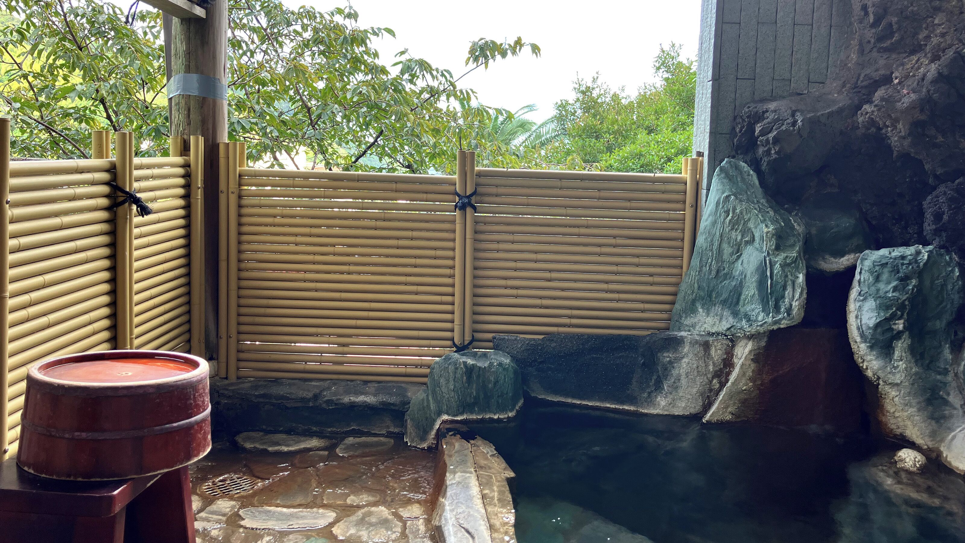 Un onsen japonés tradicional con valla de bambú, escalones de piedra y una bañera de madera.