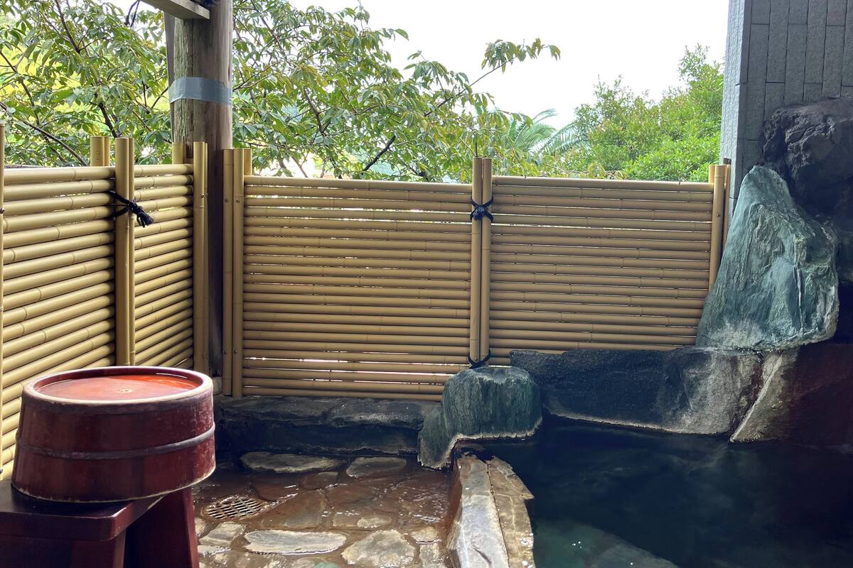 Ein traditionelles japanisches Onsen mit Bambuszaun, Steinstufen und einem Holzfass.