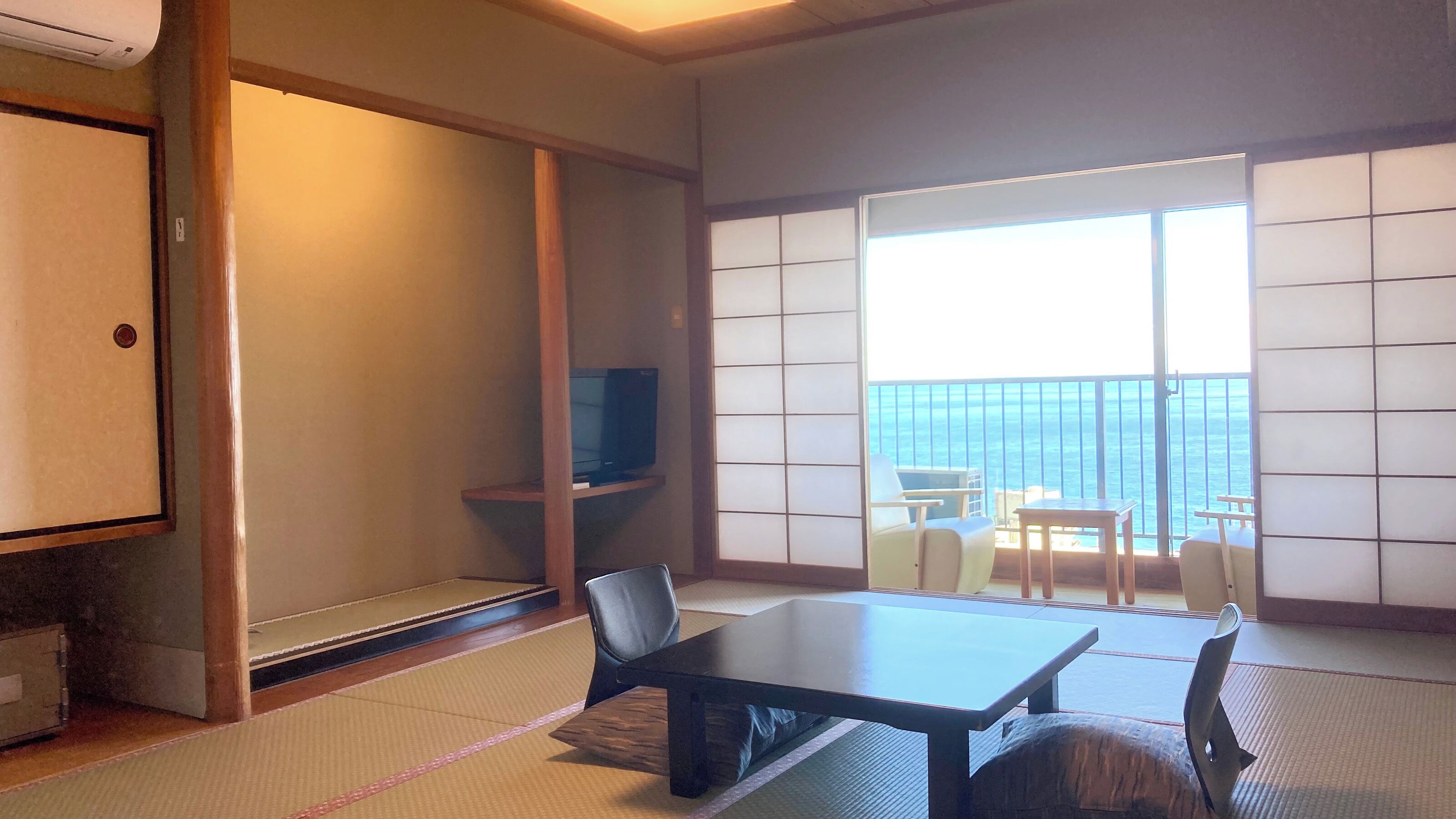 A traditional Japanese room with tatami flooring, a low table, chairs, a television, and a sliding door leading to a balcony with a view of the sea.