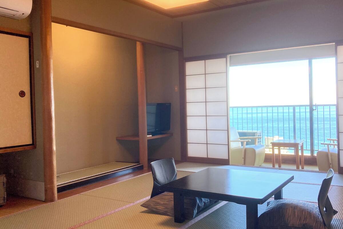 Ein traditioneller japanischer Raum mit Tatami-Boden, einem niedrigen Tisch, Stühlen, einem Fernseher und einer Schiebetür zu einem Balkon mit Meerblick.