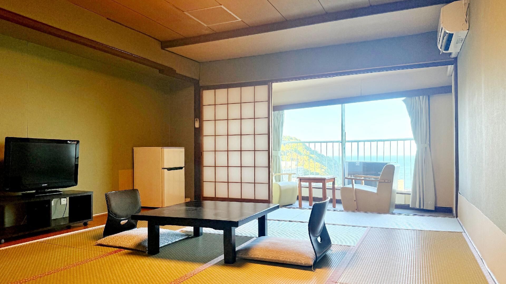 A traditional Japanese room with tatami flooring, a television, a refrigerator, and a sliding door leading to a balcony with a view.