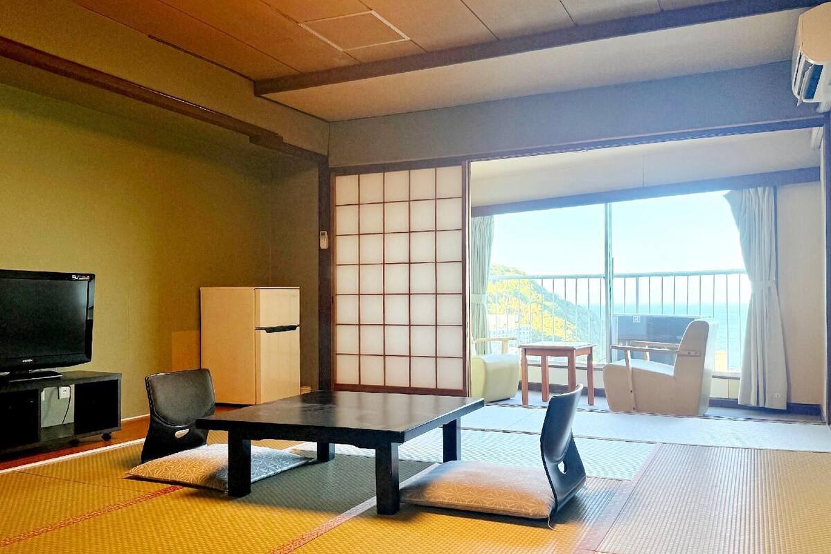 Ein traditioneller japanischer Raum mit Tatami-Böden, einem Fernseher, einem Kühlschrank und einer Schiebetür zu einem Balkon mit Aussicht.