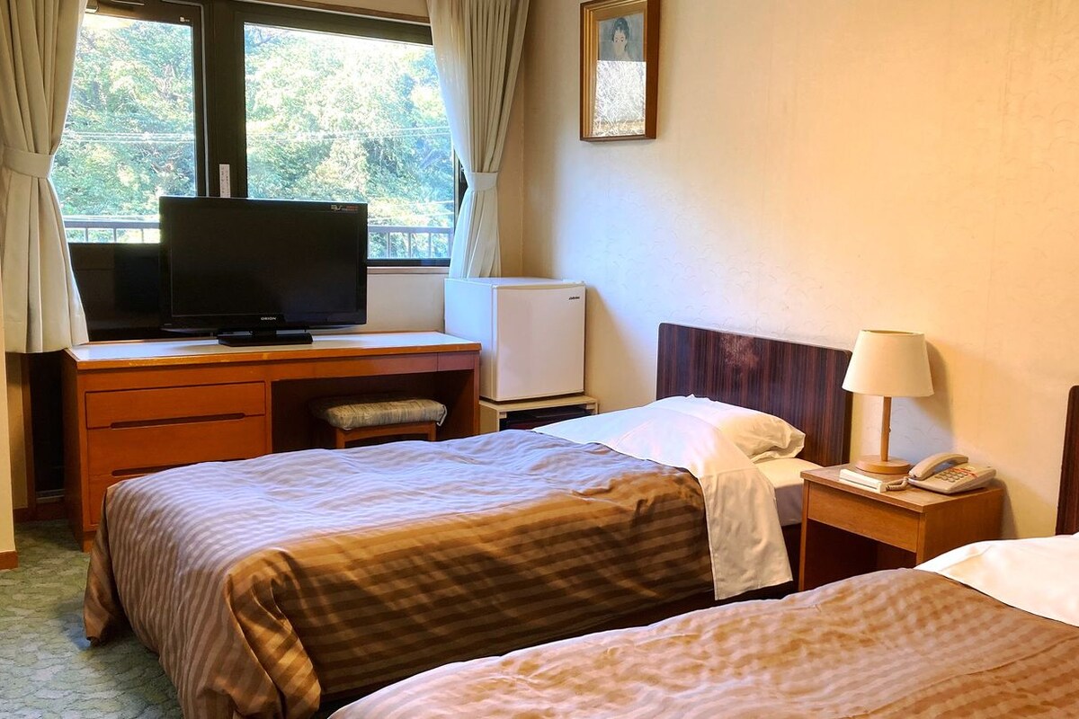 A hotel room with two beds, a desk with a television, a small refrigerator, a bedside lamp, and a telephone.