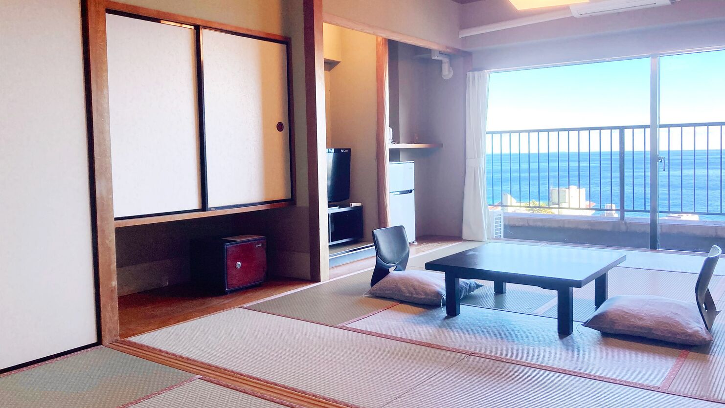 A room with tatami flooring, a low table, two cushions, a TV, and a view of the sea.