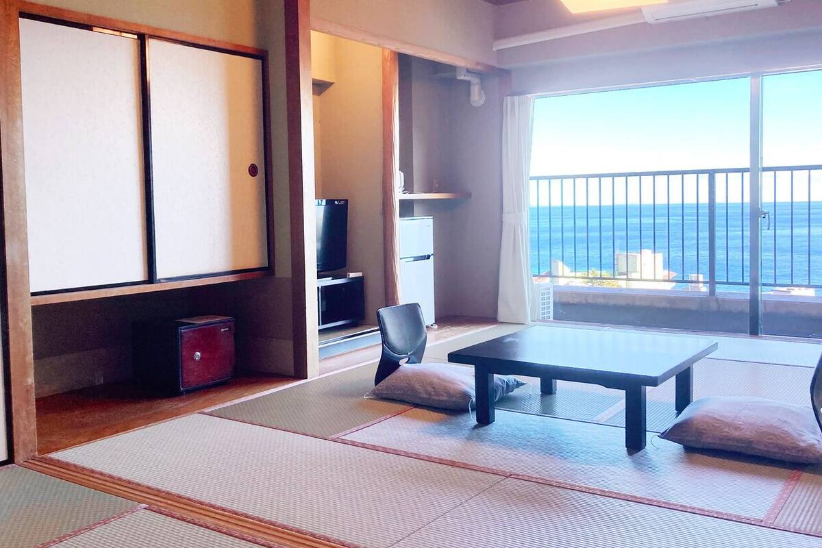 Ein Zimmer mit Tatami-Boden, einem niedrigen Tisch, zwei Kissen, einem Fernseher und Meerblick.
