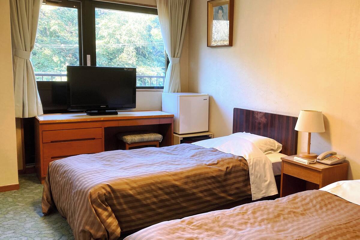 Ein Hotelzimmer mit zwei Betten, einem Schreibtisch mit Fernseher, einem Kühlschrank, einem Nachttisch mit Lampe und Telefon sowie einem Fenster mit Vorhängen.