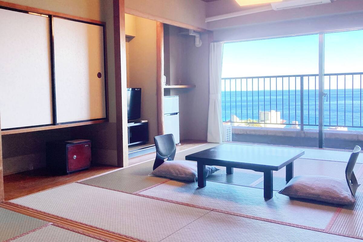 Ein Zimmer mit Tatami-Böden, einem niedrigen Tisch mit Kissen, einem Fernseher und Meerblick vom Balkon.