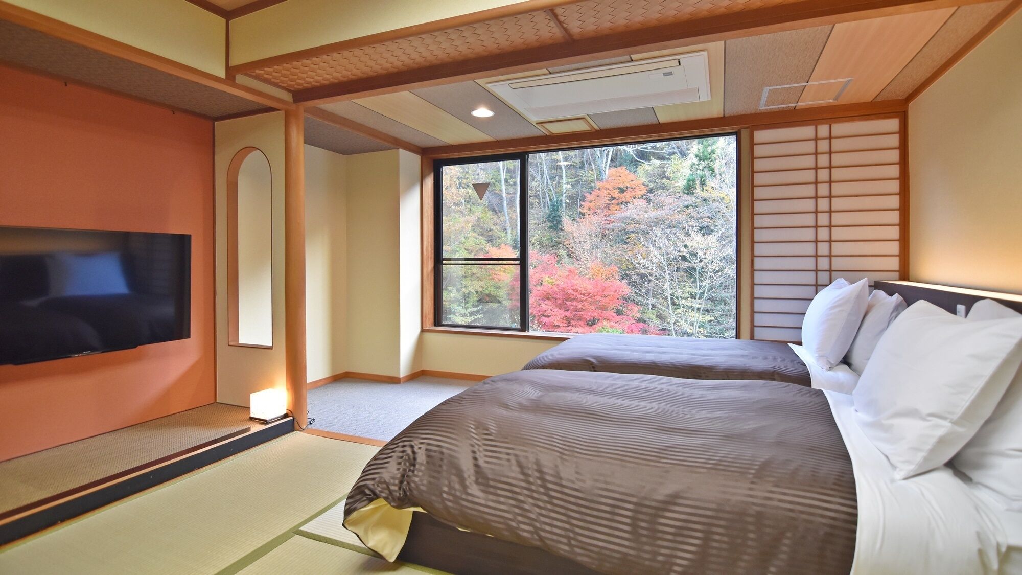 A hotel room with tatami flooring, a bed with white bedding, a television on the wall, and a large window with autumn foliage view.