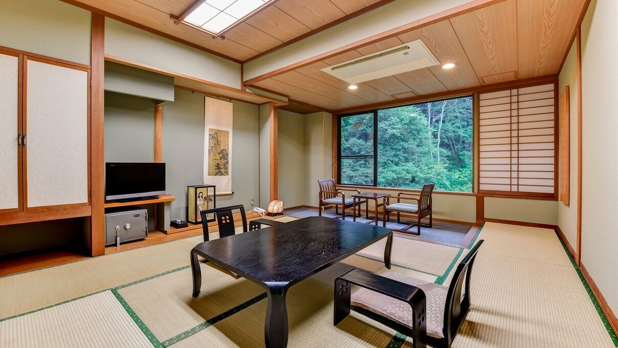 A traditional Japanese room with tatami flooring, a low table, chairs, a television, and a sliding door with a forest view.