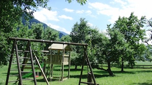 Eine Holzschaukel mit einem kleinen Spielhaus in einer grünen Wiese, im Hintergrund Bäume und Berge.