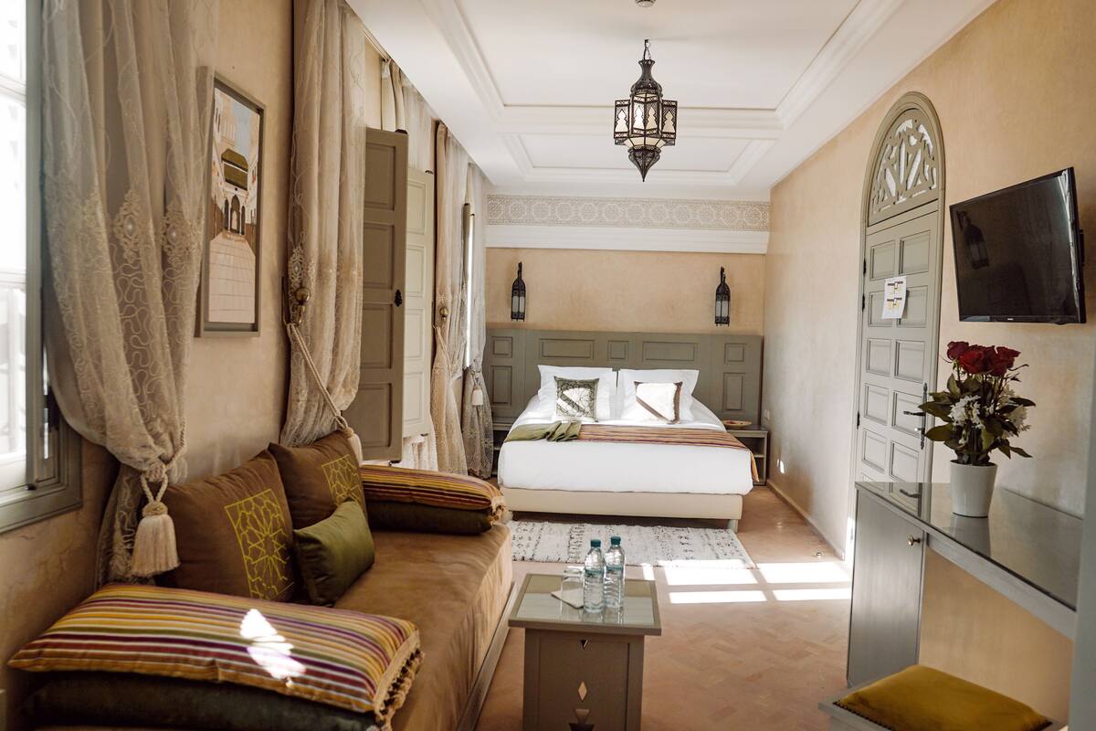 A hotel room with a bed, a sofa, a small table with water bottles, a TV, and a decorative plant.