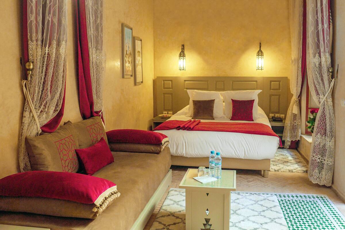 A hotel room with a bed, a sofa, a nightstand, and a small table with a bottle and glasses.