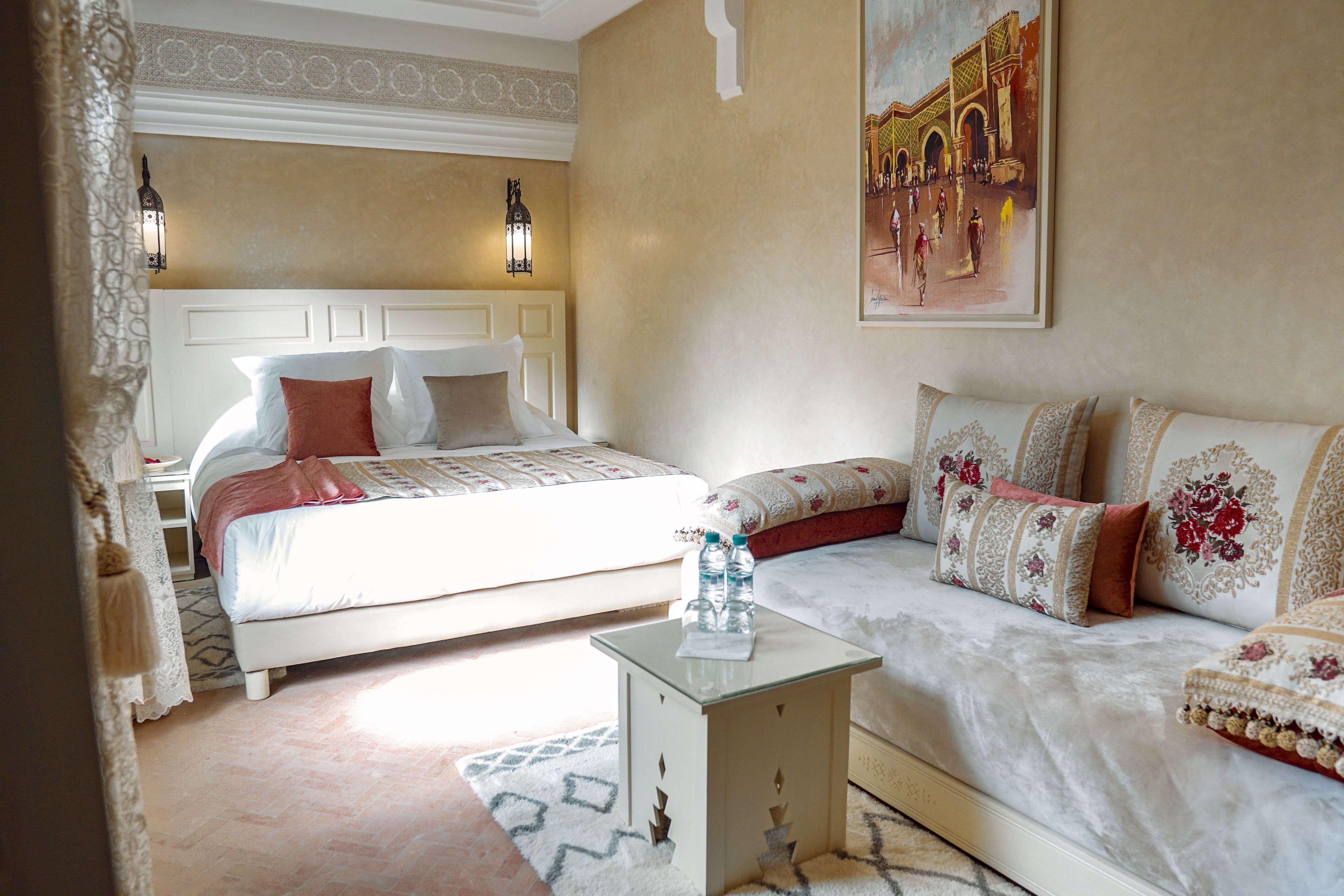 A hotel room with a bed, a sofa, a nightstand with a water bottle, and a painting on the wall.