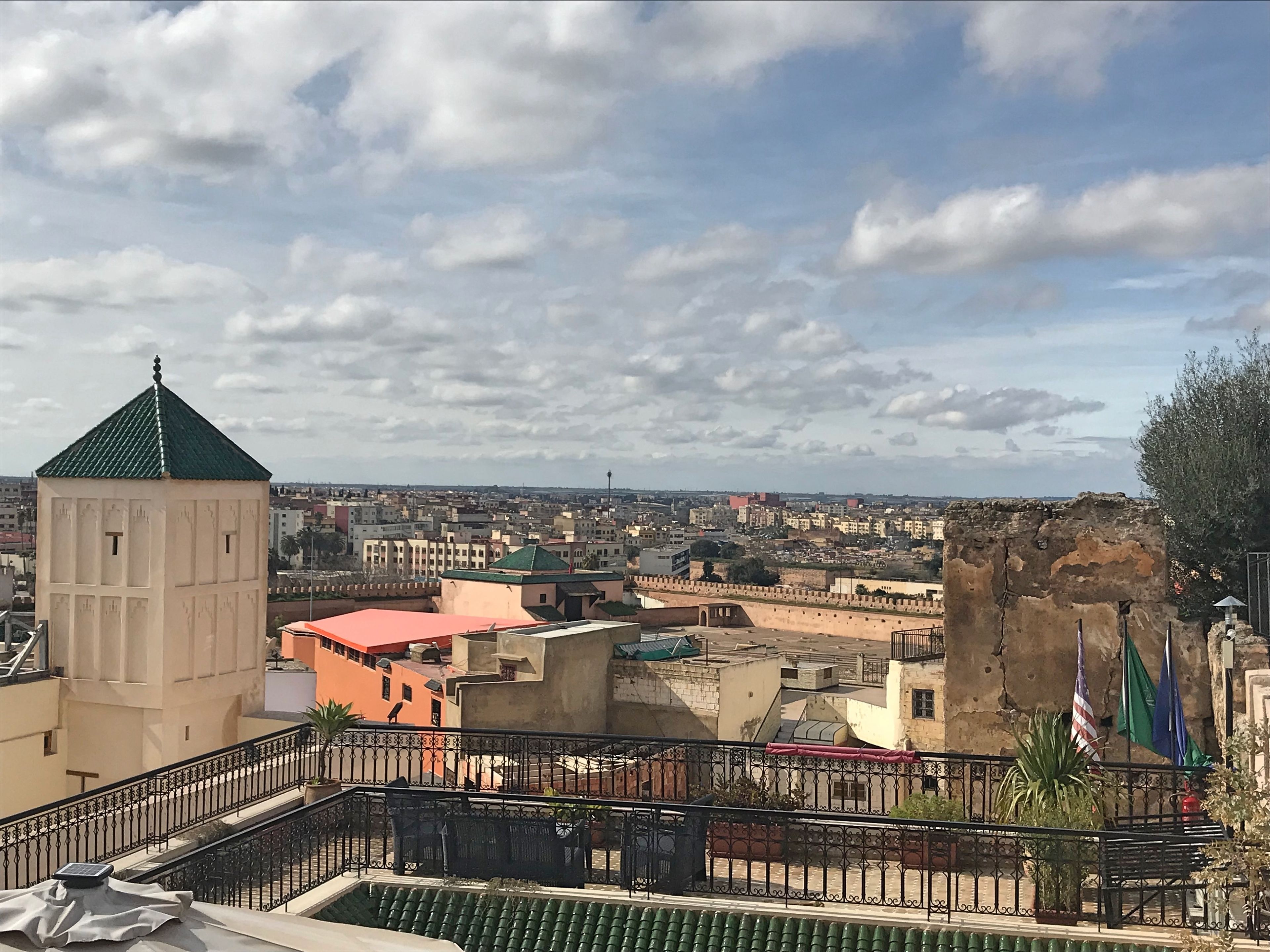 Una azotea con terraza de tejas verdes, un edificio blanco con techo verde y vistas a un paisaje urbano con diversos edificios y banderas.