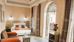A hotel room with a bed, a sofa, a small table, a nightstand, and a view of the outside balcony. - Riad Zidania (Meknes)