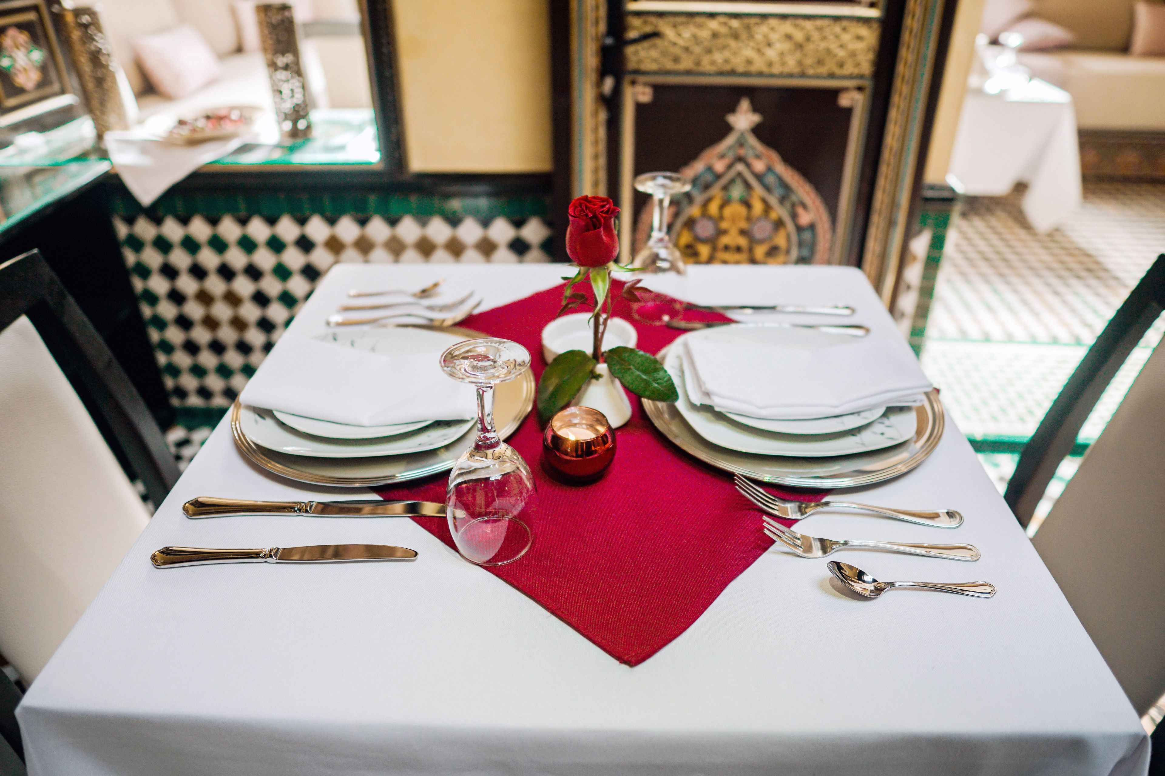 Mesa para dos con mantel blanco, servilletas rojas y una sola rosa roja.