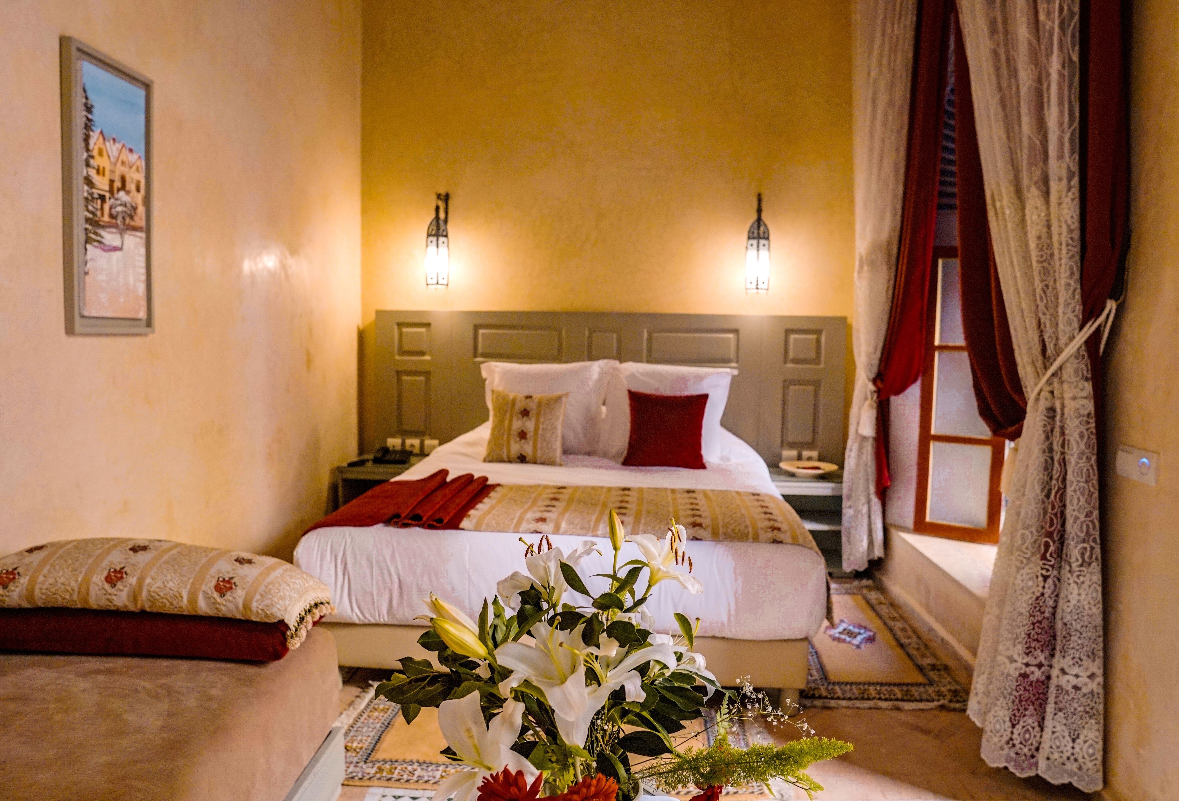A hotel room with a bed, bedside tables, a nightstand, a vase with flowers, and a window with curtains.