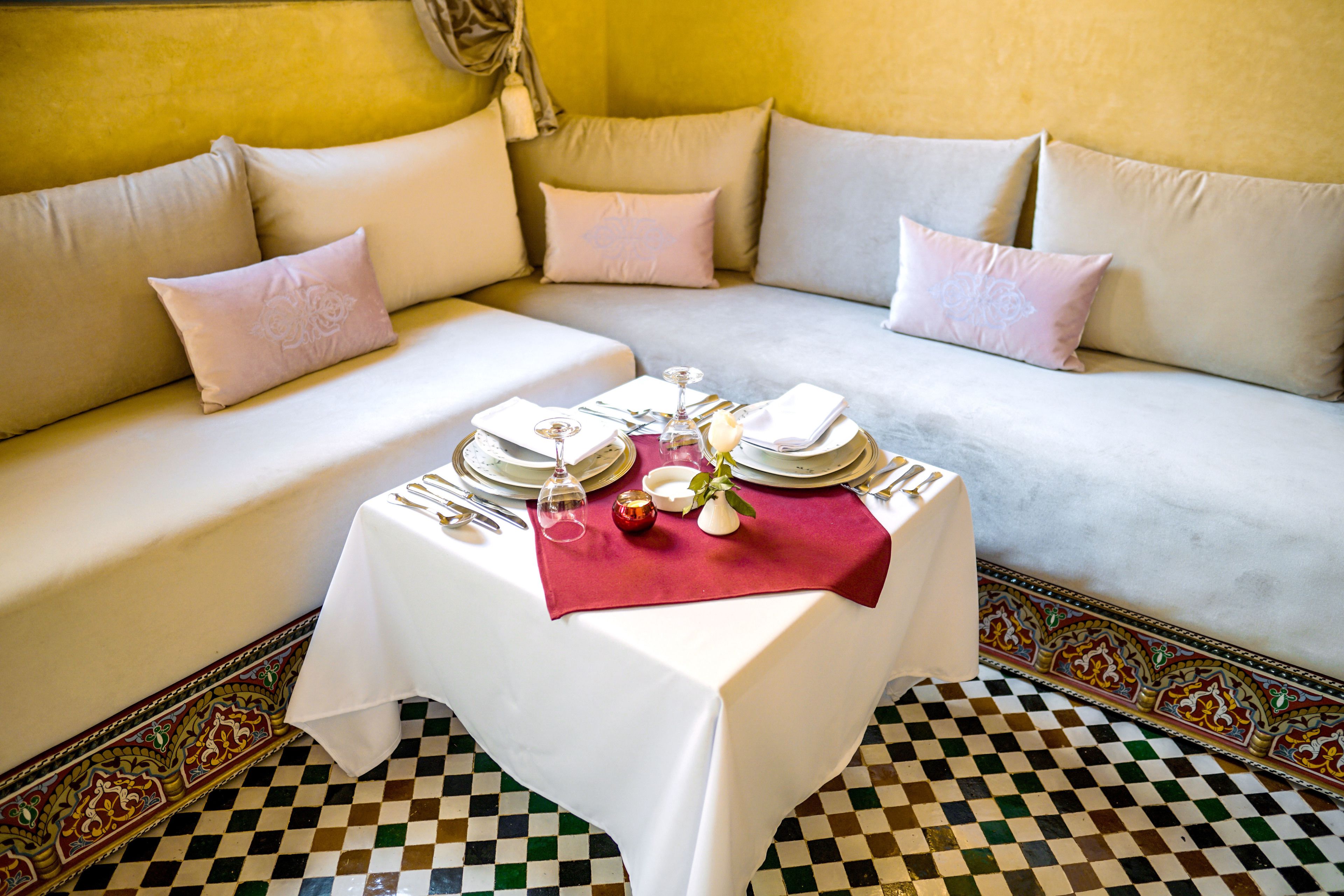 Mesa de comedor dispuesta con mantel blanco, platos, vasos y cubiertos en una habitación con suelo de cuadros y un sofá con cojines.