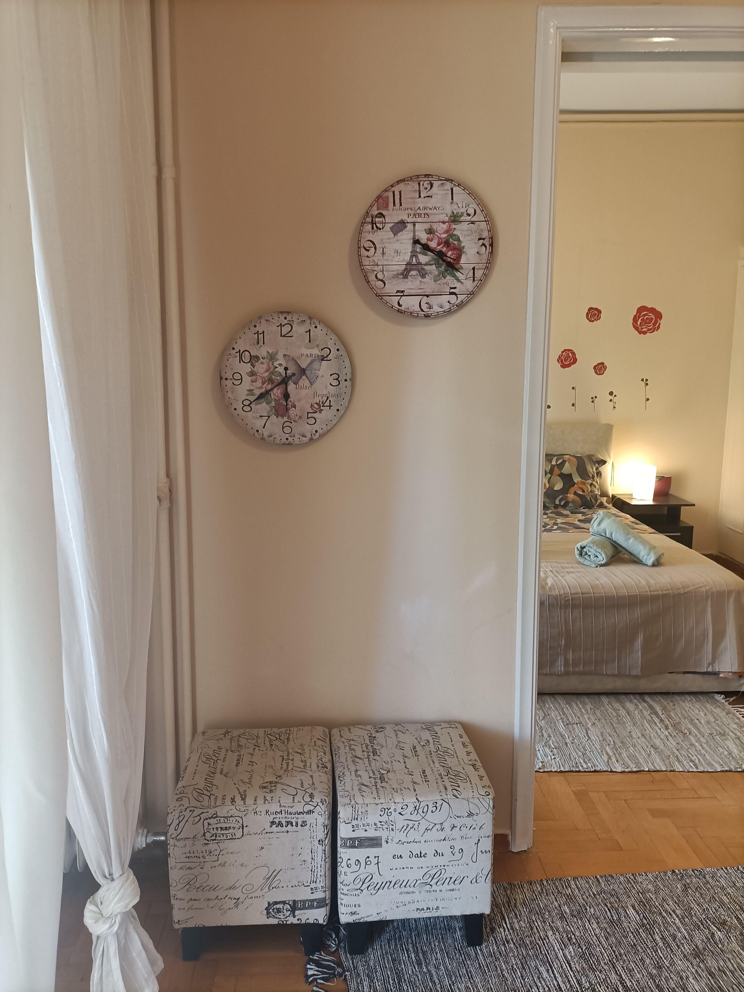 Una habitación con dos relojes en la pared, una cama con colcha estampada y una mesita de noche con lámpara.