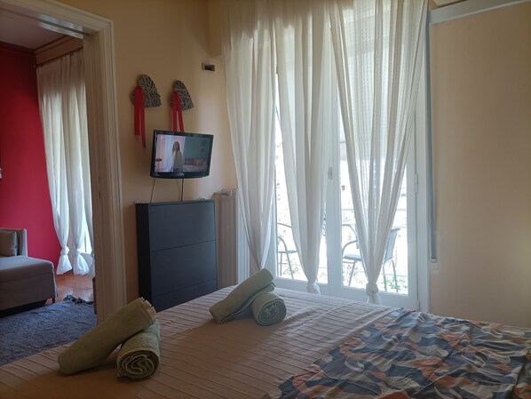 A hotel room with a bed, a television on a stand, a red wall, and a window with sheer curtains.