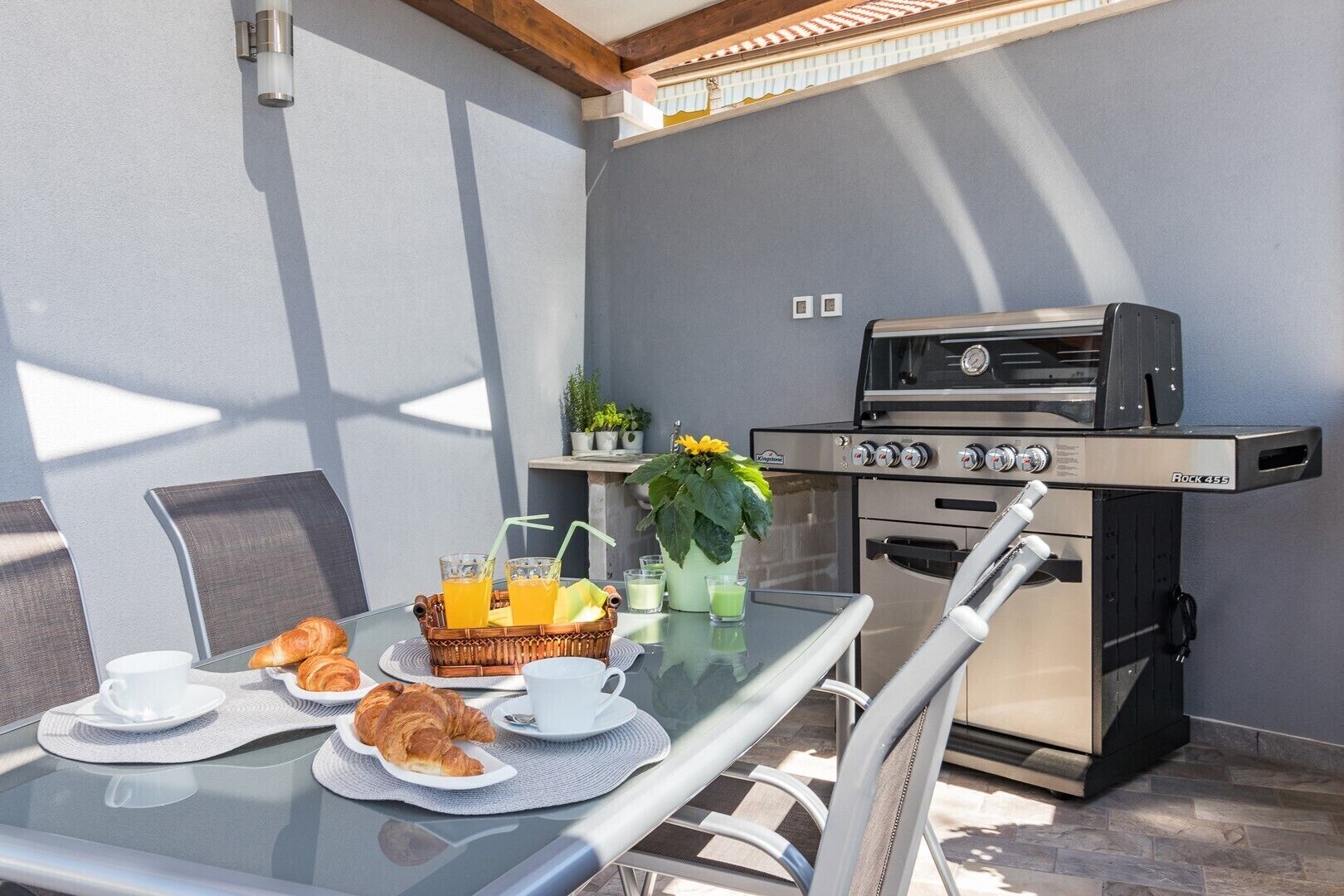 Un espace repas comprenant une table en verre, des chaises, un barbecue et un petit coin cuisine.